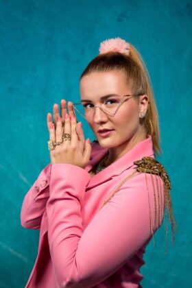 Billie Steirisch im pinken Blazer vor türkisfarbenem Hintergrund
