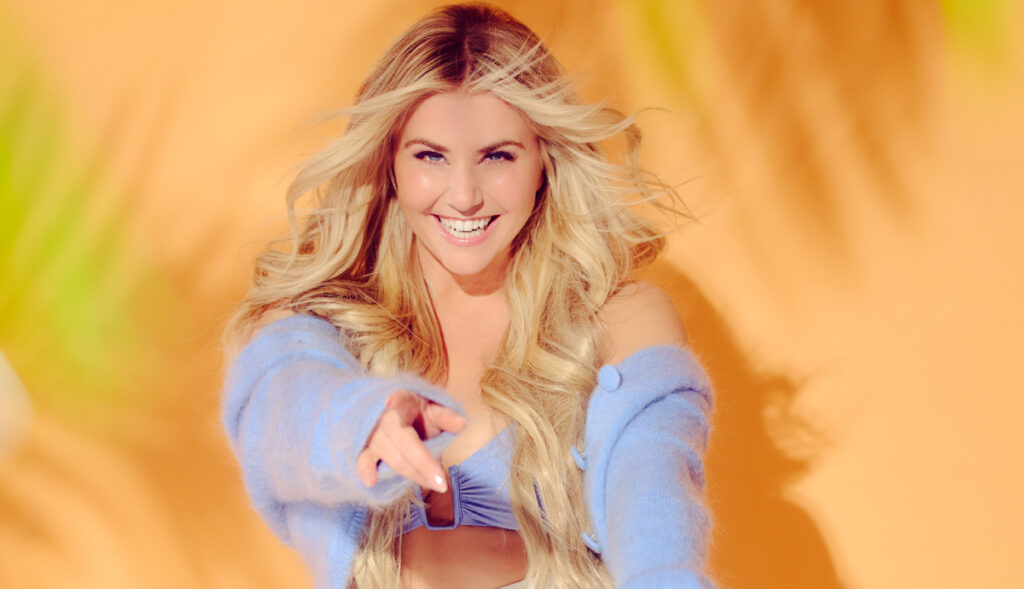 Beatrice Egli lächelt in einem hellblauen Oberteil vor orangefarbenem Hintergrund.