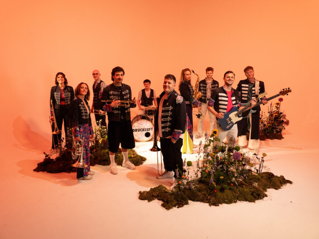 Die Band Druckluft mit Posaune, Trompete, Saxofon, Schlagzeug und Gitarre in Uniformjacken vor orangefarbenem Hintergrund