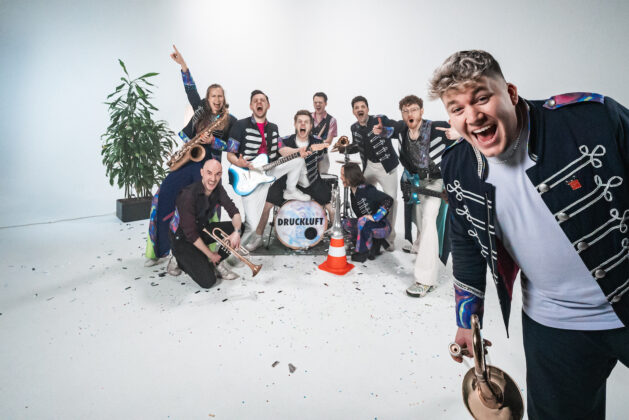 Die Band Druckluft jubelt mit ihren Instrumenten im weißen Studio, im Vordergrund ein Posaunist