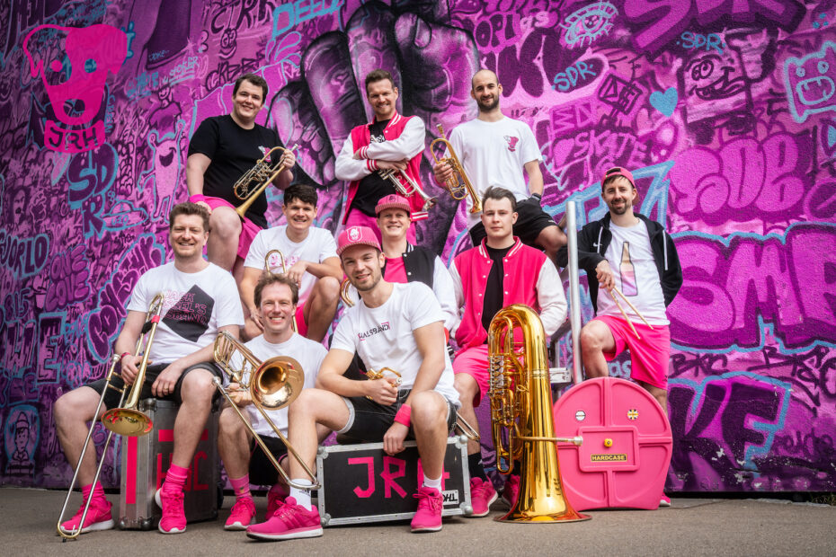 Zehn Musiker mit Trompeten, Posaunen und Tuba vor einer pinkfarbenen Graffitiwand