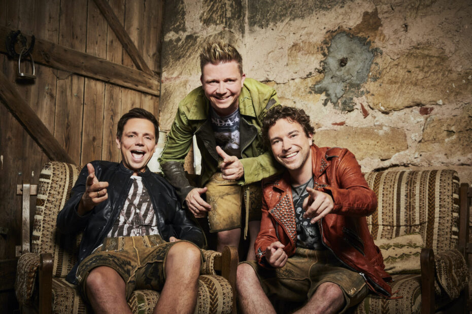 Die drei Dorfrocker in Lederhosen und Lederjacken sitzen auf Sofas vor einer alten Steinwand
