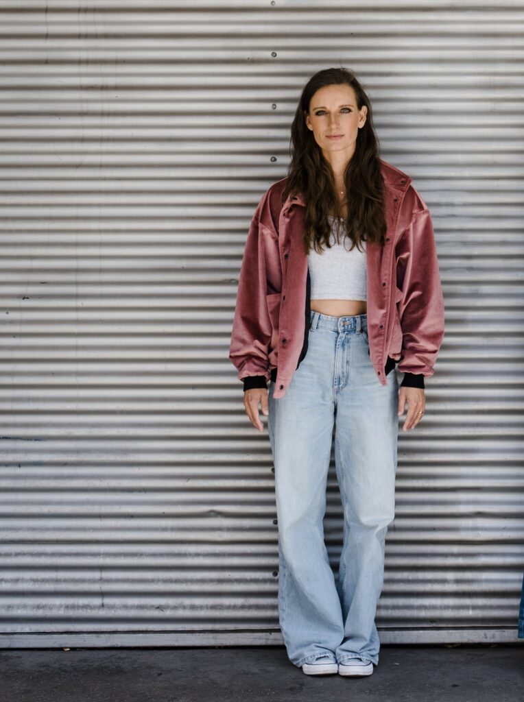 Die Mayerin in einer rosafarbenen Jacke und heller Jeans vor einer Metallwand