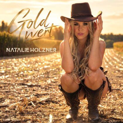 Single-Cover „Gold Wert“ von Natalie Holzner mit schwarzem Cowboyhut und schwarzem Outfit vor einer goldenen Landschaft im Abendlicht.