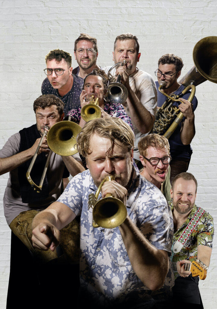 Neun Musiker von LaBrassBanda mit Trompeten, Posaune, Tuba und Gitarre vor einer weißen Ziegelwand, der Trompeter im Vordergrund zeigt direkt in die Kamera.