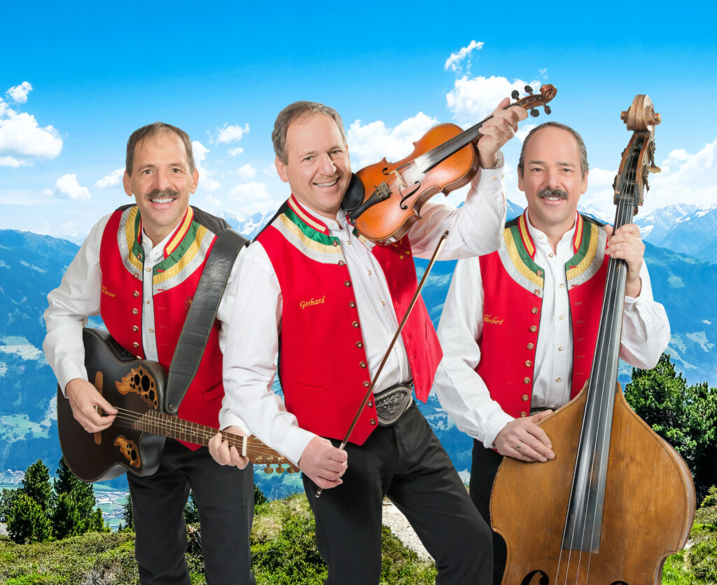 Zellberg Buam mit Gitarre, Geige und Kontrabass vor einer alpinen Berglandschaft