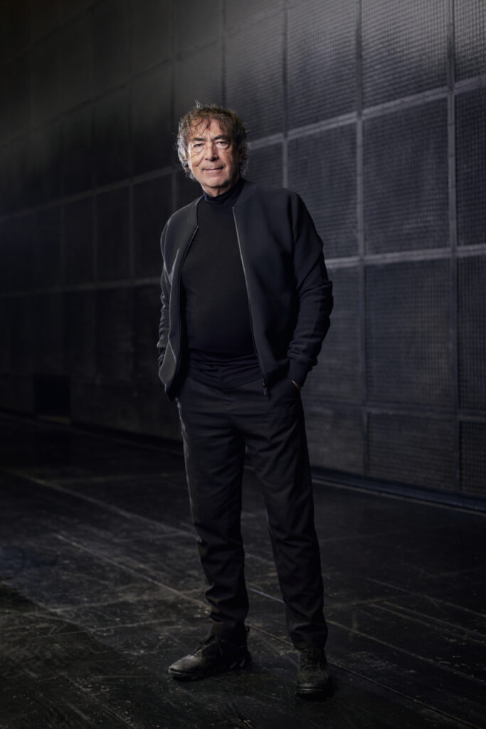 Hubert von Goisern steht mit den Händen in den Taschen in einem dunklen Raum, er trägt eine schwarze Jacke und einen Rollkragenpullover