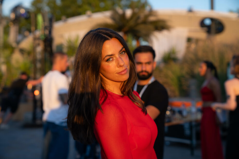 Vanessa Mai in rotem Oberteil blickt im goldenen Abendlicht über die Schulter in die Kamera, im Hintergrund sind unscharf mehrere Personen zu sehen