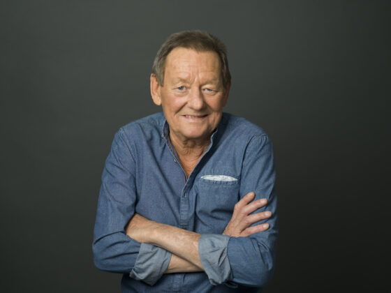 Wolfgang Ambros im blauen Hemd vor dunklem Hintergrund